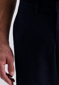 Die linke Hand eines Erwachsenen liegt entspannt neben der Taille der marineblauen, strukturierten Hose, wobei der Rand der Tasche auf weißem Hintergrund sichtbar ist.