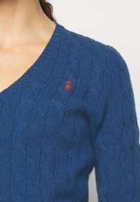 Blue cable-knit sweater made of soft yarn, featuring a V-neck design and a small embroidered logo in red on the left chest area.