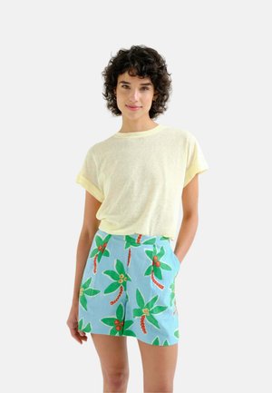 Mujer con el pelo corto y rizado que lleva una camiseta de manga corta de color amarillo claro y pantalones cortos azules con estampados de hojas tropicales verdes y rojas.