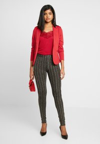 Blazer vermelho sobre uma blusa vermelha com acabamentos em renda, combinado com calças justas pretas com riscas douradas. Acessórios incluem uma pequena mala vermelha e saltos altos pretos.