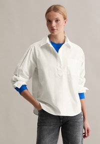 Witte katoenen shirt met een kraag, lange mouwen opgerold en een borstzak. Draagt over een blauw geribbeld top en gecombineerd met donkergrijze jeans.