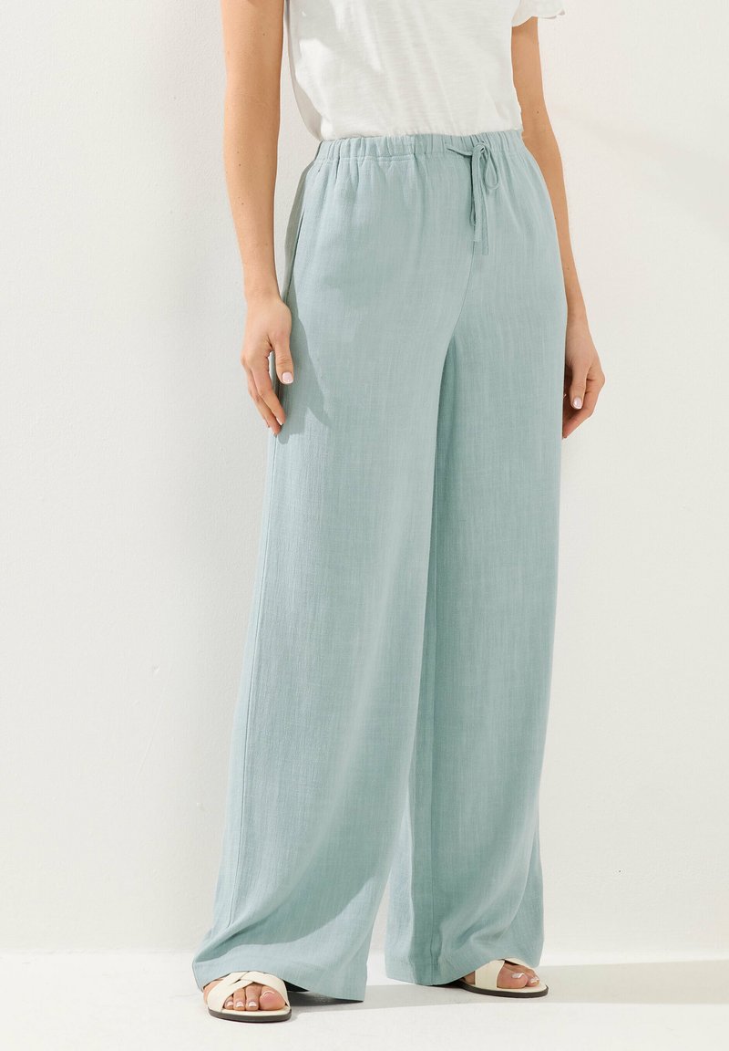 Hellblaue Leinenhose mit weitem Bein, elastischem Bund und Bindeband, kombiniert mit weißem Oberteil und weißen offenen Sandalen vor weißem Hintergrund.