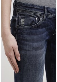 Jeans de mezclilla índigo con forma ajustada, que presentan un pequeño bolsillo frontal etiquetado como "GS RAW 520" y remaches metálicos. La textura es ligeramente desgastada.