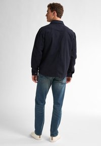 Chemise en coton marine à manches longues, avec poignets boutonnés et coupe droite ; associée à un jean en denim bleu déchiré et des baskets beiges avec des accents.
