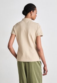Beige poloskjorte med korte ermer og en tettsittende design, laget av glatt bomullsstoff. Kombinert med løse grønne bukser, med sidespalter synlige.