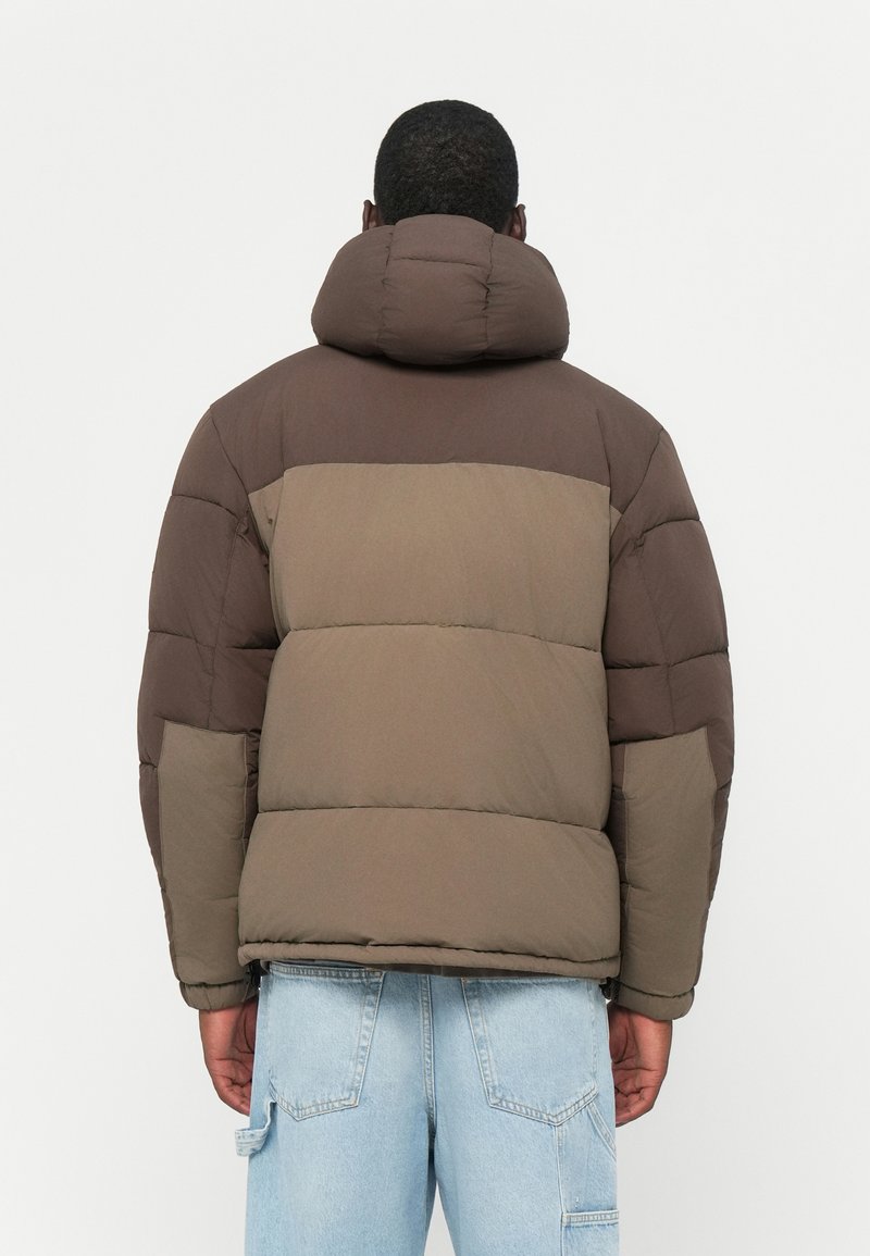 Chaqueta acolchada marrón con secciones acolchadas, con un alto cuello y capucha. La tela tiene un acabado mate. La vista trasera muestra un bolsillo de parche en la manga.