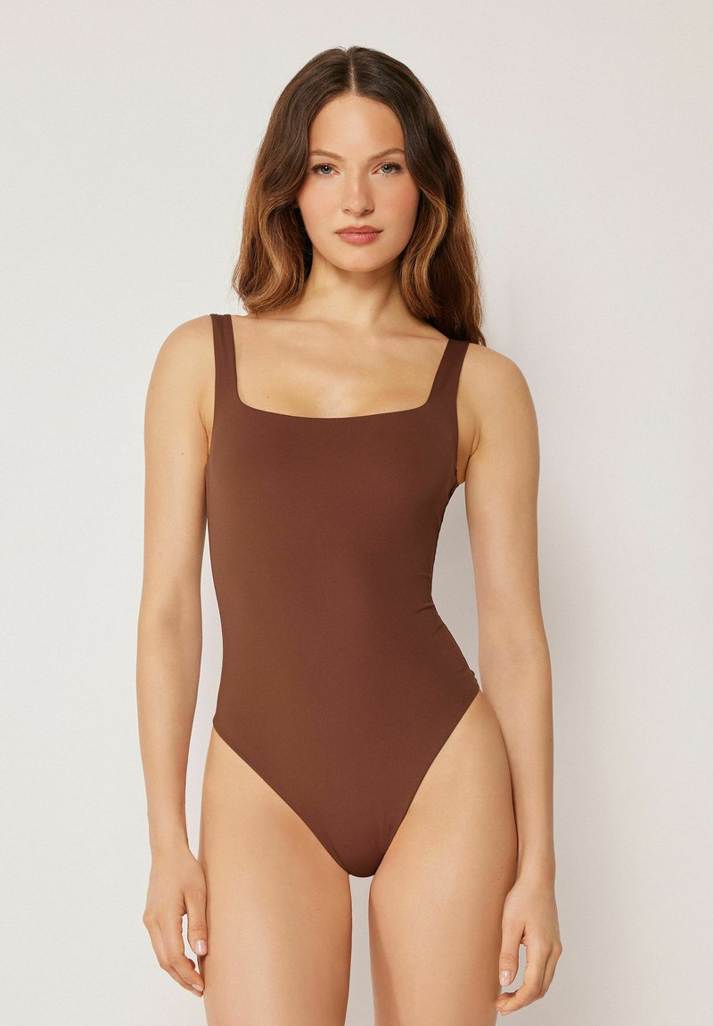 Femme aux longs cheveux ondulés portant un maillot de bain une-pièce marron uni, sans manches, à encolure carrée, debout devant un fond clair uni.