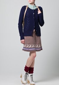 Cardigan en tricot marine avec un design en câble, fermeture à boutons ; chemise vert clair ; jupe brune à motifs de renne ; bottines blanches à lacets.