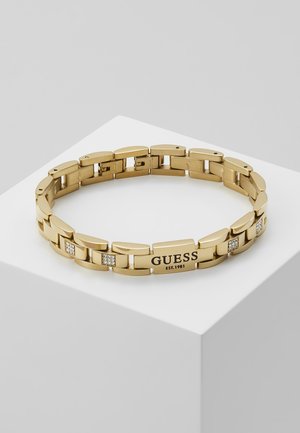 Bracelet en chaîne métallique ton doré avec petits cristaux et plaque gravée "GUESS EST. 1981", présenté sur un socle blanc.