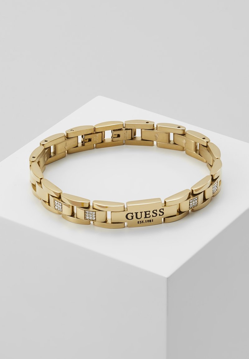Bracelet en chaîne métallique ton doré avec petits cristaux et plaque gravée "GUESS EST. 1981", présenté sur un socle blanc.