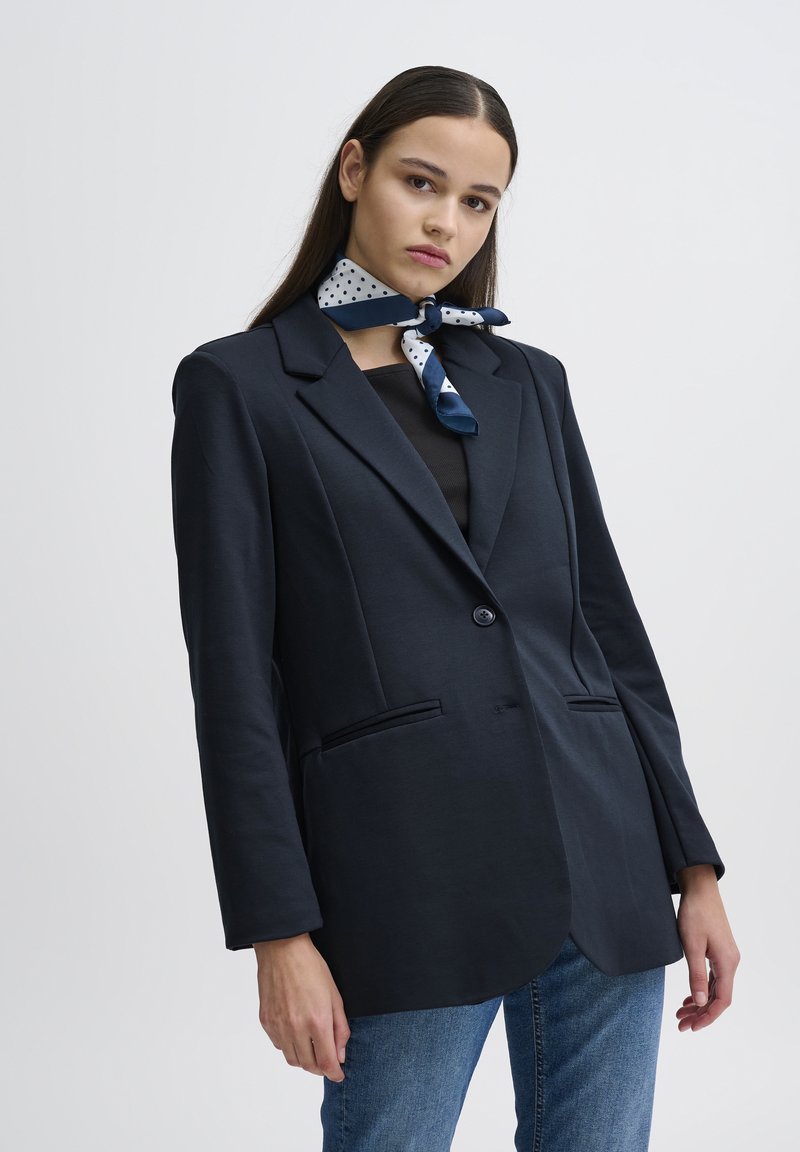 Navy blazer o dopasowanym kroju, z dwoma przednimi kieszeniami, pojedynczym zapięciem na guzik i miękkiego materiału. Połączony z chustą w kropki zawiązaną wokół szyi.