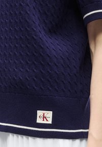 Ναυτικό πλεκτό πουλόβερ με υφασμένο σχέδιο, λευκή ρίγα στο τελείωμα και κόκκινο εμβληματικό λογότυπο Calvin Klein κοντά στη μέση στο σώμα ενός ατόμου.