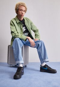 Ung man med lockigt blont hår sitter på en silverväska, iklädd en grön jacka, svart Nike-t-shirt, blå jeans, vita strumpor och svarta sneakers.