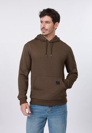 Mężczyzna w teksturowanej brązowej bluzie z kapturem i kieszenią z przodu oraz jasnoniebieskich dżinsach, patrzący lekko w lewo na jednolitym tle.