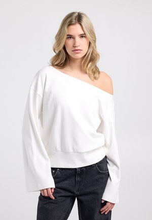 Sweat-shirt blanc à épaules dénudées avec manches larges et taille ajustée. Tissu doux avec une texture lisse. Associé à un jean foncé.