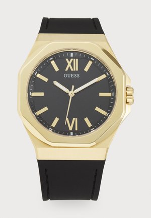 Montre à cadran noir avec de grands index en or et des chiffres romains. Présente une lunette octogonale en or et un bracelet en caoutchouc noir.