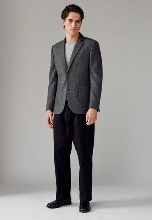 Jeune homme debout portant un blazer gris, une chemise gris clair, un pantalon noir et des chaussures noires sur un fond gris uni.