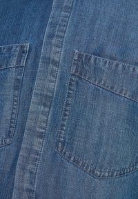Camisa de denim en un tono azul medio, con dos bolsillos en el pecho y una ligera textura visible en la tela, además de detalles de costura visibles.
