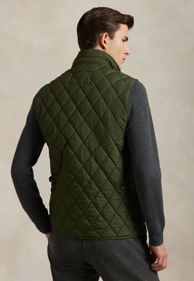 Hombre con chaleco acolchado verde oscuro sobre camisa de manga larga gris oscuro, de pie con la espalda y la cabeza giradas ligeramente hacia un lado.