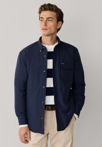 Jonge man die een marineblauwe jas met knopen draagt over een wit met marineblauw gestreepte trui, beige broek en een bruine riem, kijkt opzij.