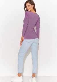 Fioletowa bluzka z długim rękawem z marszczonymi ramionami, w połączeniu z jasnoniebieskimi dżinsami i białymi sportowymi trampkami na koturnie. Bluzka ma mały złoty akcent na plecach.