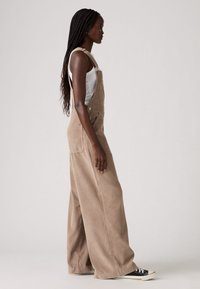 Brown corduroy overalls with wide legs, silver hardware, large front pocket, worn over a white tank top, paired with black sneakers.