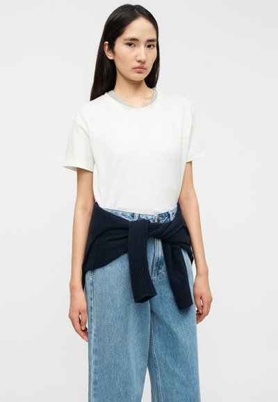 Λευκό βαμβακερό t-shirt με στρογγυλή λαιμόκοψη, συνδυασμένο με ανοιχτό μπλε παντελόνι με ευρύ πόδι. Ένα ναυτικό πουλόβερ είναι δεμένο γύρω από τη μέση.