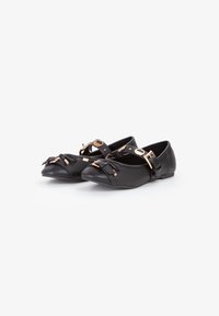 Ballerine nere a punta con cinturini alla caviglia, fiocchi decorativi e fibbie in tono oro su uno sfondo bianco.