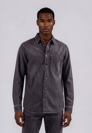 Ung mand iført en falmet sort denimskjorte med knapper og matchende bukser, stående mod en ensartet lys baggrund.