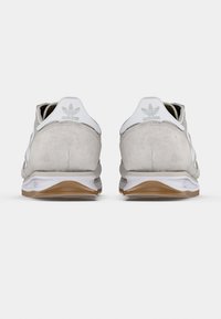 Fehér és világosszürke sportcipők velúr hatással, gumitalppal és minimalista dizájnnal. Az Adidas logó a sarkon és csíkok díszítik.