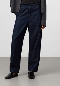 Mörkblå, högmidjade jeans med avslappnad passform. Har klassisk femfickedesign och diskret sömnad. Bärs med svarta slip-in skor.