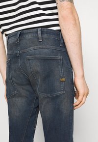 Person som bär mörkblå denimjeans med bakfickor och en liten G-Star Raw-etikett, kombinerat med en svartvit randig skjorta.