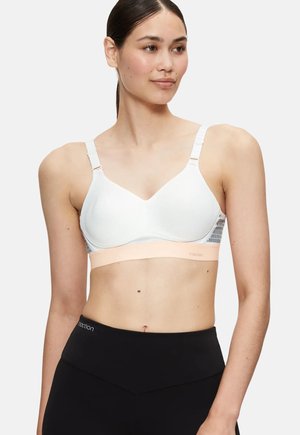 Jeune femme portant un soutien-gorge de sport blanc et des leggings noirs taille haute, regardant légèrement sur le côté avec une expression neutre.