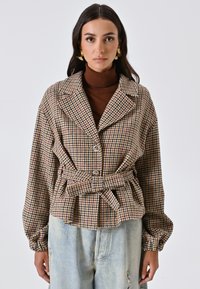 Giacca a quadri houndstooth con cintura in marrone, crema e rosso, con colletto, due bottoni e maniche voluminose. Abbinata a jeans strappati.