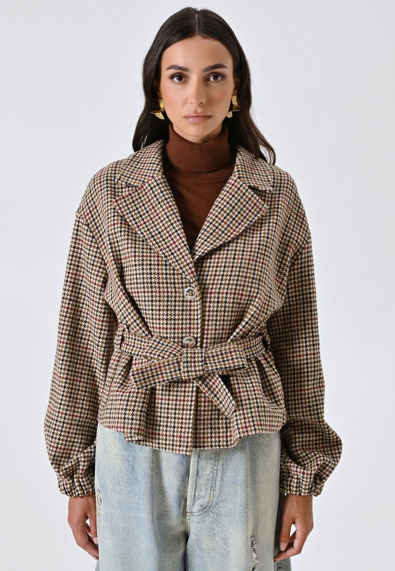 Giacca a quadri houndstooth con cintura in marrone, crema e rosso, con colletto, due bottoni e maniche voluminose. Abbinata a jeans strappati.