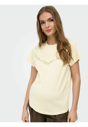 Ung kvinde med langt hår iført en cremefarvet kortærmet bluse med dekorative kvaster og brune bukser, stående mod en ensartet baggrund.