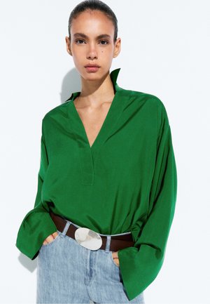 Vrouw draagt een losse groene blouse, ingestopt in lichtblauwe jeans met een donkere riem en grote ovale zilveren gesp, staand tegen een witte achtergrond.