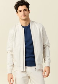 Lichtbeige bomberjack met ribgebreide manchetten, een rits aan de voorkant en een gladde textuur. Gecombineerd met een aansluitende marineblauwe trui en lichte broek.