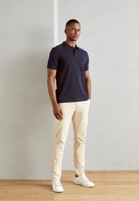 Mørkeblå polo-skjorte med korte ærmer, kombinert med lysebeige chinos og hvite sneakers. Glatt stoff og skreddersydd design. Minimalistiske detaljer.