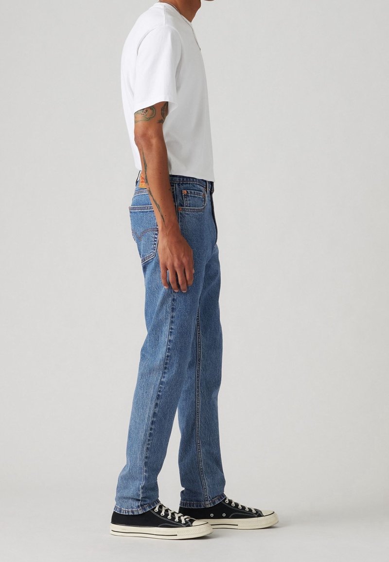 Homme portant un jean bleu clair, un t-shirt blanc à manches courtes, des baskets basses noires, debout de profil devant un fond clair uni.