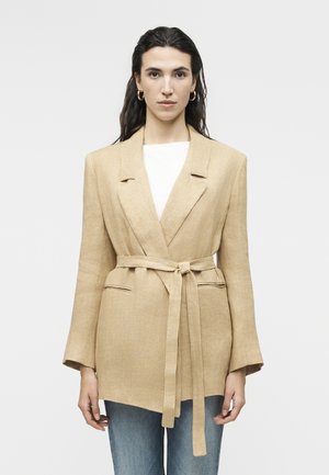 Pikajuukseline naine, kes kannab kuldseid rõngaskõrvarõngaid, beežikat vöödiga bleiserit, valget topi ja siniseid pükse, seisab ühetaustal helendava taustaga ees.