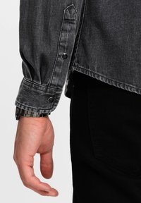 Grijze denimshirt met knoopmanchetten, toont een gestructureerde stof en zwarte drukknoopjes. Hand rust nabij de onderzoom, gecombineerd met zwarte broek.