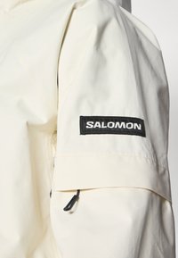 Jachetă bej deschisă, dintr-un material texturat, având un patch cu logo-ul negru "SALOMON" pe mânecă, cordon ajustabil și un design elegant.