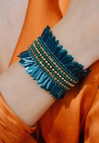 Bracelet turquoise avec des formes superposées en forme de feuilles, des perles dorées et vertes, et des accents scintillants ; porté au poignet sur un fond orangé.