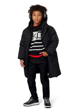 Zwarte gewatteerde jas, grijze graphic sweatshirt met zwart-witte strepen, jeans en zwarte sneakers met grijze accenten. Koptelefoon op.