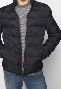 Black puffer jacket with quilted design and zip closure, worn over a patterned grey sweater. Casual fit with elastic cuffs and hem.