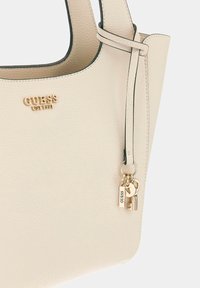 Crème kleur tote bag van pebble leer met donkere afwerking, voorzien van gouden logo, sleutelhanger en hangslot accent. Korte handgreep voor gemakkelijk dragen.