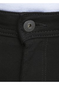Primer plano de la cintura de unos pantalones vaqueros de mezclilla negra con un botón de metal visible y una tela blanca por encima de la cinturilla.