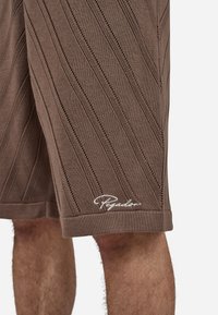 Brune strikkede shorts har diagonalt ribbestruktur, kontrasterende små hull og et hvitt logodetalj ved kanten. Designet for komfort og pusteevne.
