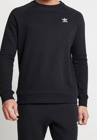 Černá mikina s dlouhými raglánovými rukávy, kulatým výstřihem a bílým logem Adidas na hrudi. Měkká látka, žebrované manžety a lem.